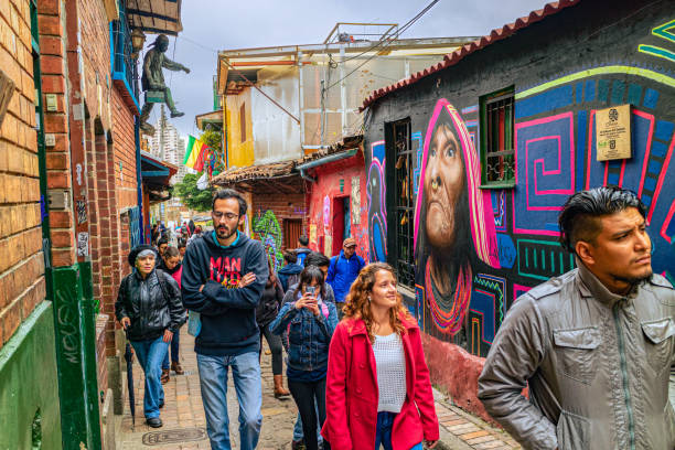 Bogota, Colombia - Tourists And Local Colombians On The Calle del Embudo, In The Historic La Candelaria District Of The Andes Capital City In South America. Bogota, Colombia - July 20, 2016: Both Tourists and local Colombian people walk up the narrow Calle del Embudo one of the most colorful streets in the historic La Candelaria district of Bogotá, the Andean capital city of the South American country of Colombia. The street leads to the Chorro de Quevedo, the plaza where it is believed the Spanish Conquistador, Gonzalo Jiménez de Quesada founded the city in 1538. Many street facing walls in this area are painted with either street art or the legends of the pre-Colombian era, in the vibrant colours of Colombia. The altitude at street level is 8,660 feet above mean sea level. Photo shot on an overcast morning; horizontal format. calle del embudo stock pictures, royalty-free photos & images