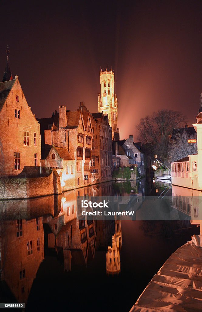 Cold night in Bruges Belgium Night shot of the belfry in Bruges with reflection of the surrounding historic buildings in the water. The belfry is surrounded by fog. Baroque Style Stock Photo