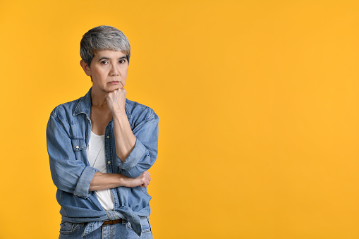 Image of worried, thinking middle aged Asian woman 50s standing isolated over colour background, looking at camera