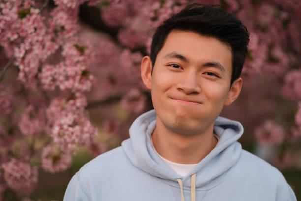 lovely cute Asian young man making face, looking at camera. Blur pink sakura flowers background