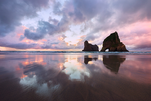 A dramatic and colourful sunset is reflected in the sand at Wharariki Beach on the South Island of New Zealand. The Archway Islands are silhouetted against the sky.