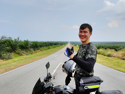 An Asian man is sending thumbs up while feeling accomplished riding motorcycle on road trip.