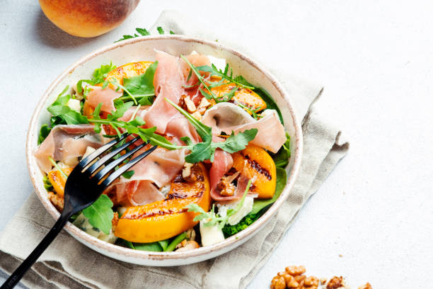 Summer salad bowl with sweet grilled peach, jamon, soft cheese, walnuts and fresh arugula on white kitchen table background, top view, negative space Summer salad bowl with sweet grilled peach, jamon, soft cheese, walnuts and fresh arugula on white kitchen table background, top view, negative space prosciutto stock pictures, royalty-free photos & images