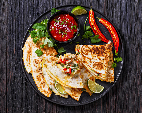 mexican quesadilla with chicken, green onion, cheese, chilli pepper, coriander leaves filling  and tomato salsa on black plate on dark wood table, flat lay, close-up