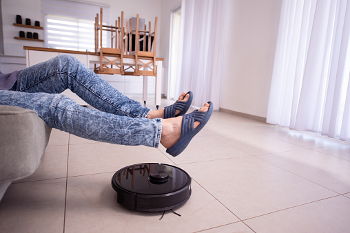 Close-up image of female's legs raised up as the robot vacuum passes and cleans the living room's tiled floor. The inconvenience of removing obstacles when a robotic vacuum cleaner working. New problems of modern life with home automation.