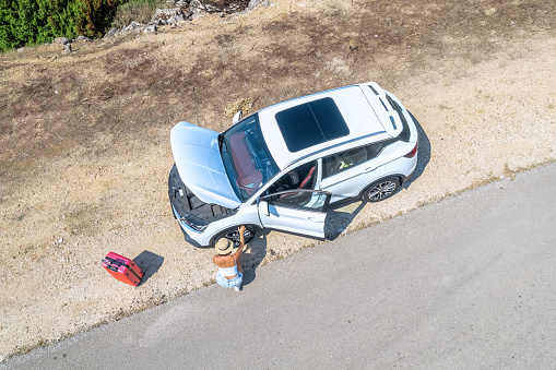 Woman sitting by broken car with open hood and checking  wheel at countryside road. Aerial, top, drone view