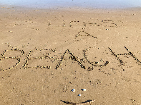 The words 'Life’s a Beach' have been written in big letters in the sand, with a shell as a full-stop. In front of the words is a smiley face, which has been made out of three shells and some driftwood. It is an idyllic and peaceful scene on a sunny day.