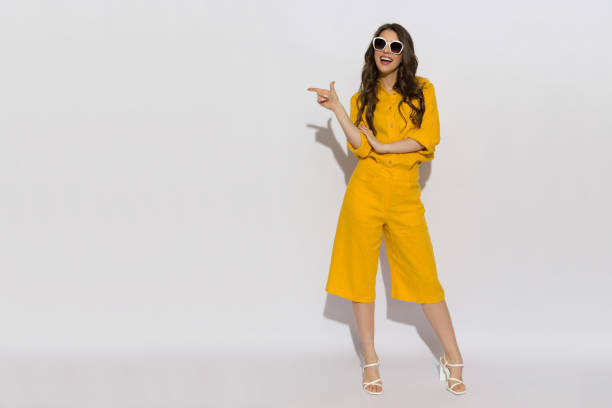 Young woman in linen shirt, shorts and high heels pointing to the side and talking. Beautiful young woman in sunglasses, yellow linen shirt and shorts standing, pointing to the side and talking. Full length studio shot. whole stock pictures, royalty-free photos & images