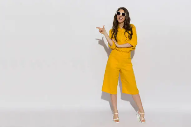 Beautiful young woman in sunglasses, yellow linen shirt and shorts standing, pointing to the side and talking. Full length studio shot.