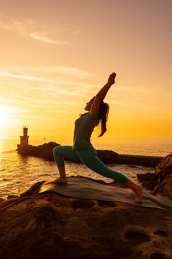 Anjaneyasana, A woman doing meditation and yoga exercises on a rock at sunset next to a lighthouse in the sea, healthy and naturist life