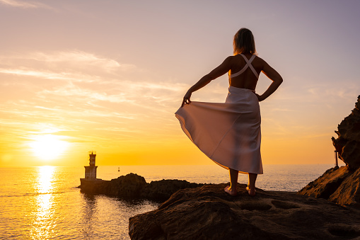 A blonde woman in a white dress at sunset next to a lighthouse in the sea, healthy and naturist life, lifestyle, looking at the sea