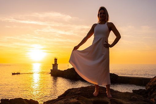 Portrait of a blonde woman in a white dress at sunset next to a lighthouse in the sea, healthy and naturist life, lifestyle