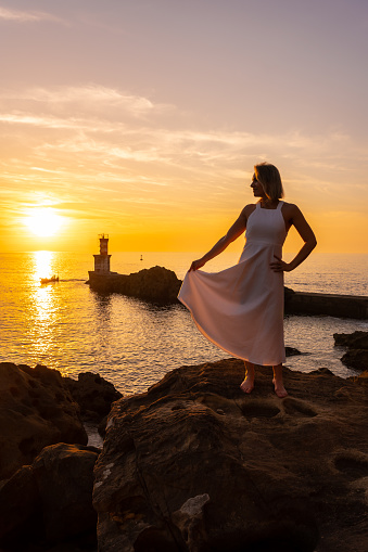 A blonde woman in a white dress at sunset next to a lighthouse in the sea, healthy and naturist life, lifestyle