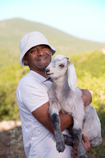 Portrait of a farmer wearing a white bucket hat, holding a kid goat, standing outdoors with a scenic view of green hills.
