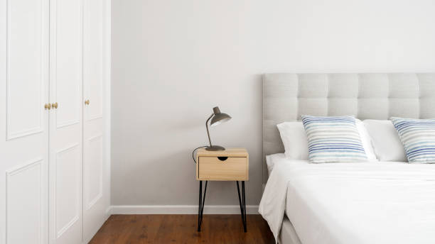 Side table with lamp standing close to bed and wardrobe Bedroom interior design in cozy modern house. Web banner view of electric light lamp stand on wooden side table close to bed and wardrobe. Apartment with comfortable furniture queen size bed stock pictures, royalty-free photos & images