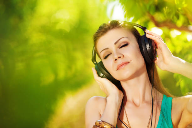 Young attractive woman in a green tank top enjoys and relaxes listening to light music in big black headphones relaxing alone in a summer green park..Beautiful student girl listening to music on a smartphone via headphones while relaxing outdoors.