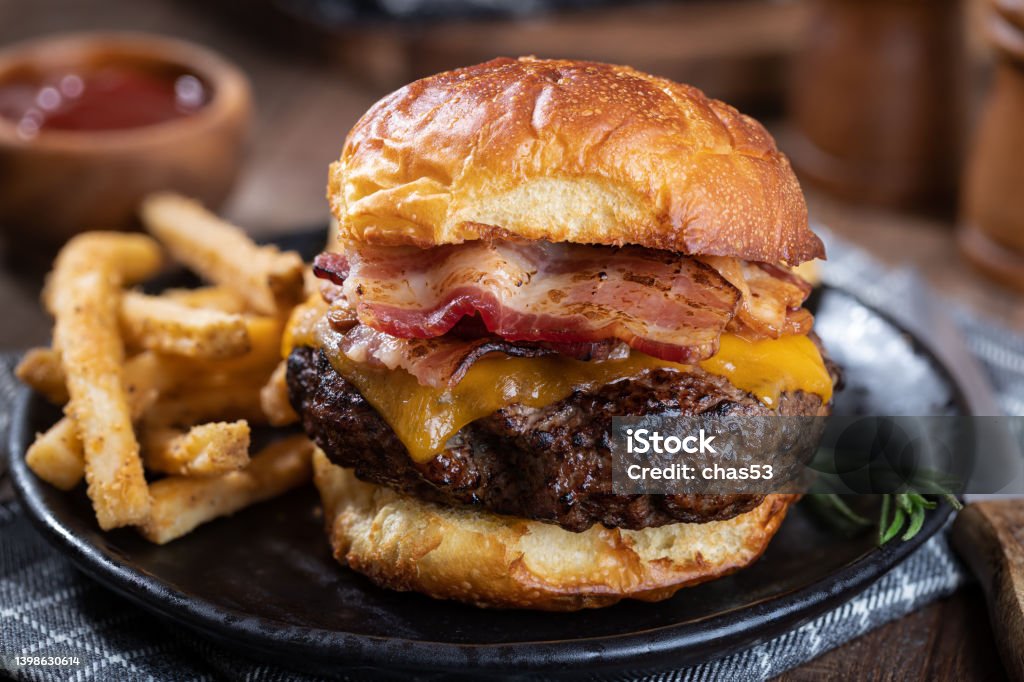Bacon cheeseburger on a toasted bun Closeup of a bacon cheeseburger on a toasted bun and french fries on a black plate Burger Stock Photo