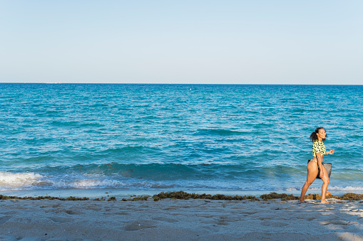 Beautiful Latin brunette walks along the beach while carrying her laptop and communication equipment in her bag to continue working while resting