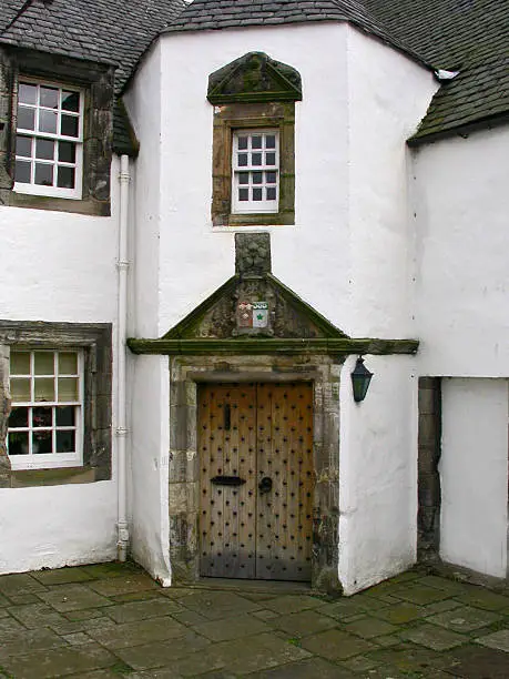 Iron studded old wooden door set in stone pillaster and lintol doorframe. With stone surround window frames to the left, set in white painted walls.Stone paving stones in the foreground.