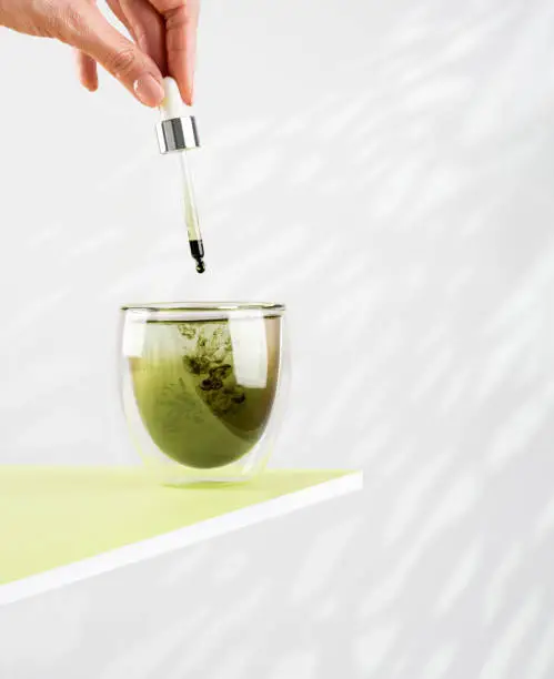 Female hand pours liquid chlorophyll into a glass of water with a dropper. Glass of liquid chlorophyl on the green table. White background, natural sunlight. Concept of superfood, healthy drink