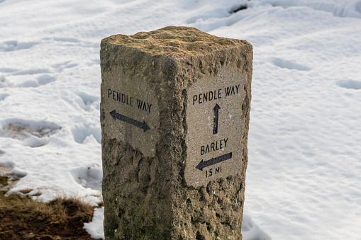 Snow and footprints around stone waymarker on the Pendle Way hiking trail at the top of Pendle Hill in Lancashire, England.