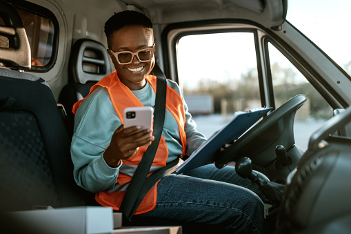 Young smiling female delivery driver texting while working, she is inside of a delivery van