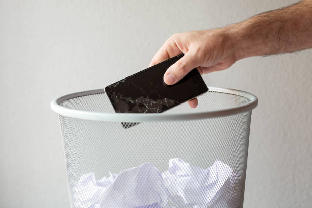 A man's hand throws a broken mobile phone in the trash on a gray background.