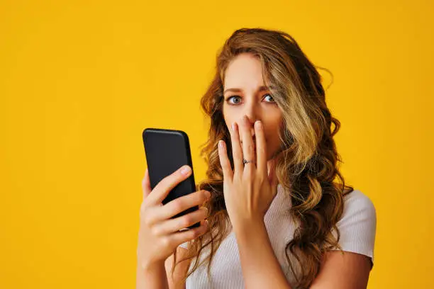 young attractive woman with smartphone cell phone long hair wearing a white t-shirt with shocked surprised face smiling and looking to the camera studio