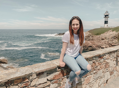 Young brown-haired woman, happy and smiling, sitting by a lighthouse facing the sea. Summer concept, holidays, tourism.