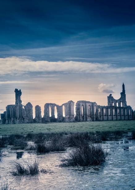 Medieval ruins of Byland Abbey set against a dark dusk sky with the setting sun shining upon the waterlogged marshland in the foreground.