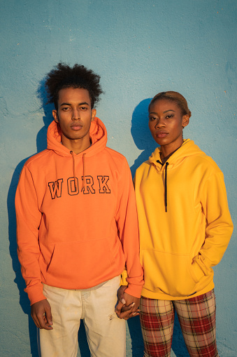 Studio shot of a couple holding hands, dressed in brightly colored sweatshirts and staring at the camera against a blue wall