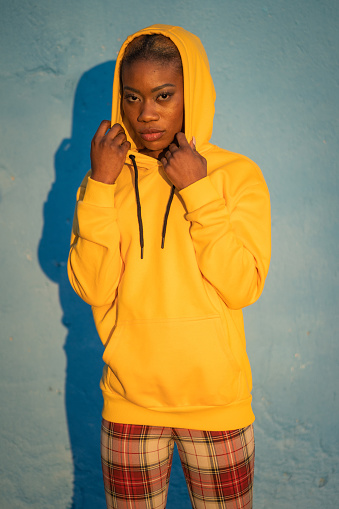 An Afro-Caribbean woman is staring at the camera, holding the hood of her yellow sweatshirt with both hands and covering her head, her shadow can be seen against the blue background