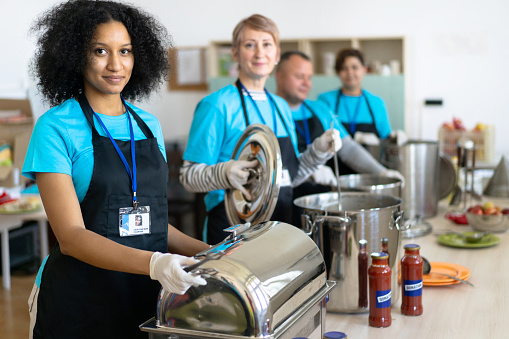 Multiethnic volunteers waiting to serve homeless people with hot food from their serving pans, wearing aprons and matching t-shirts and identity lanyards