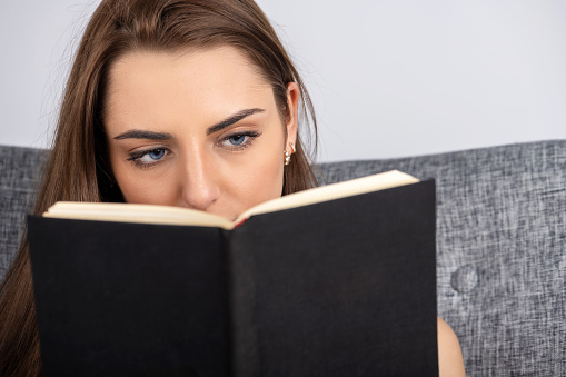 Leisure and hobbies concept. Portrait of beautiful brunette woman with blue eyes reading book. Book with blank black old and aged cover and copy space. Model eyes looking to book