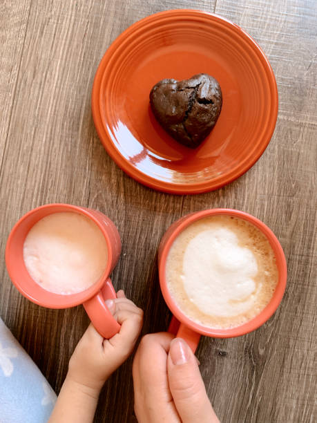 A Mother & 3-Year-Old Daughter's Hands Holding Matching Pink Mugs Filled with Hot Milk Lattes and Sharing a Heart-Shaped Chocolate Muffin