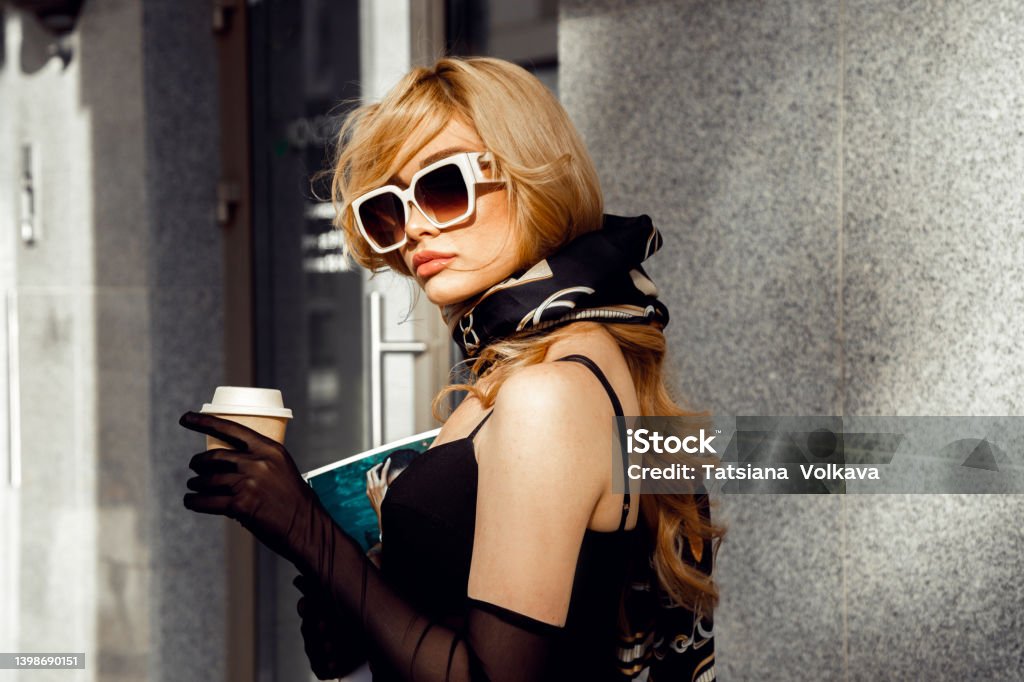 Side view of young pretty woman with long wavy fair hair wearing black dress, long mesh gloves, silk printed kerchief. Side view of young pretty woman with long wavy fair hair wearing black dress, long mesh gloves, silk printed kerchief, white sunglasses, standing near building in city, holding coffee in takeaway cup. Coffee - Drink Stock Photo