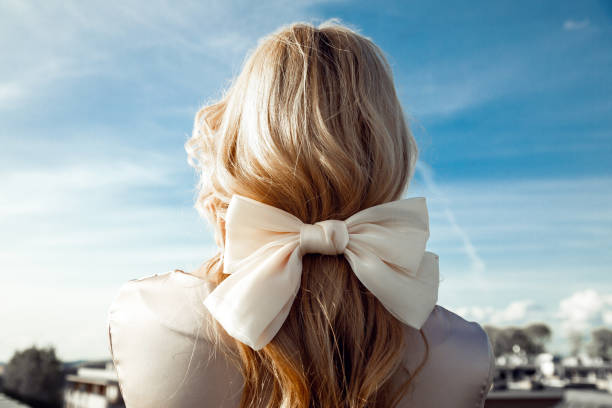 vista trasera de la mujer con el pelo largo y rubio decorado con un lazo beige hecho de cinta de pie sobre el fondo azul del cielo. primer plano - bow building fotografías e imágenes de stock