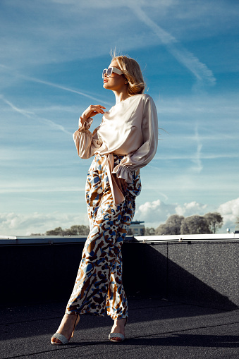 Portrait of young confident woman with long fair hair wearing beige long-sleeve silk blouse, printed trousers, high-heeled shoes, sunglasses, standing with crossed legs on roof, posing at sunset.