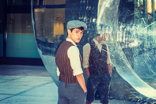 City Boy. Wearing newsboy cap, light yellow shirt, patterned vest, Asian American college student standing by metal mirror wall in New York, turning back, looking at you.
