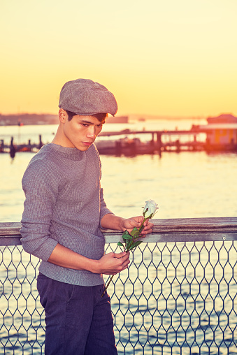 Asian American college student seeking love in New York, wearing newsboy cap, knitting sweater, holding white rose, standing by river in sunset, looking down, sad, thinking.