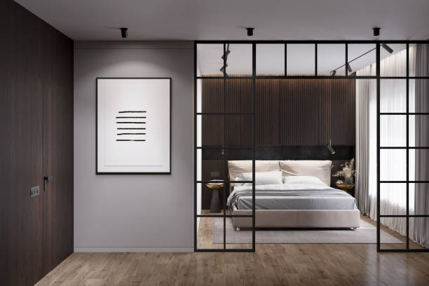 Modern dark interior with the illuminated vertical poster on a gray wall between a wooden door and a glass partition overlooking a modern bedroom with a light bed by the window. Front view.