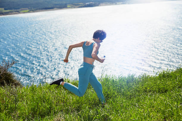 eine junge sportliche frau, die an sonnigen sommertagen in der natur über die hügel an der seebucht joggt - running track women running spring stock-fotos und bilder