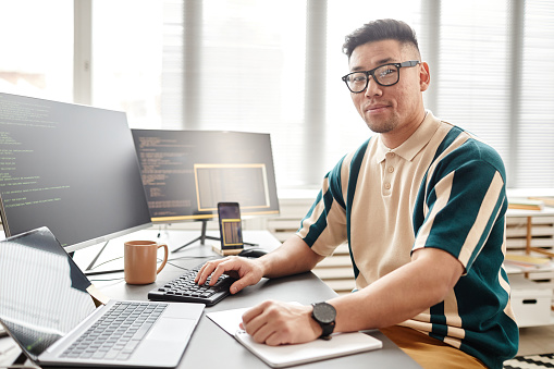 Warm toned portrait of Asian man as IT developer or QA engineer smiling at camera while sitting at workplace with computers, copy space