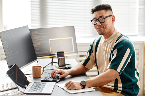 Warm toned portrait of Asian IT developer or QA engineer looking at camera while sitting at workplace with computers, copy space