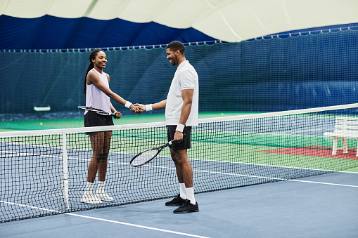 Full length shot of two tennis players shaking hands across net during match at indoor court, copy space