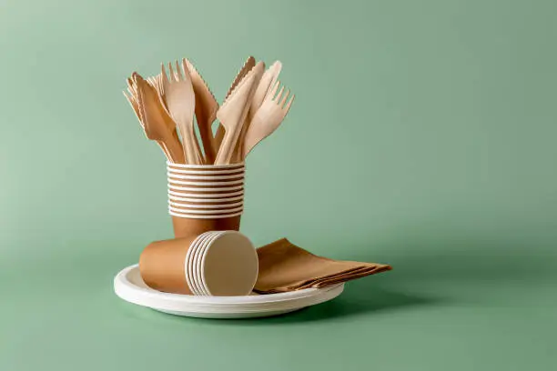 Photo of Stack of eco-friendly disposable tableware. Wooden forks and knives, paper cups and plates against green background. Biodegradable cutlery and dishes for picnics, takeaways. Copy space.