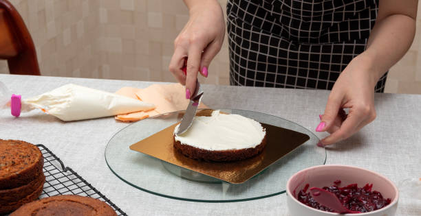 Leveling the cream on the sponge cake with spatula. There is bowl with stuffing on the table. Next to it is pastry bag with cream, and clean biscuits. The process of making cake. Selective focus. Picture for articles about food, confectioners. stock photo