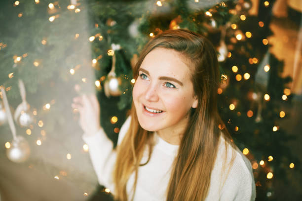 Young pretty girl looking up throught the window, christmas tree on background stock photo