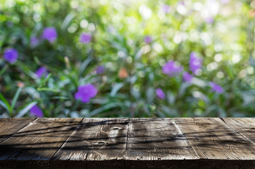 Empty wooden table with shadow of trees in outdoor. Summer or spring background with green juicy foliage and purple flowers. Natural template with bokeh and sunlight for product display on top of the table. Focus on foreground.