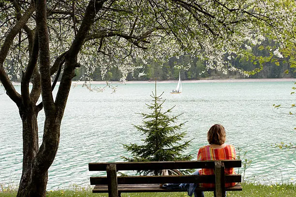 A woman is sitting on a bench, looking to the little sailing boat. Walchensee, Bavaria.